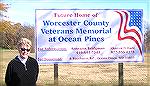 Roseann Bridgman, co-chairman of the Worcester County Veterans Memorial at Ocean Pines fund raising committee, poses next to the sign announcing the proposed memorial near the Ocean Pines South Gate P